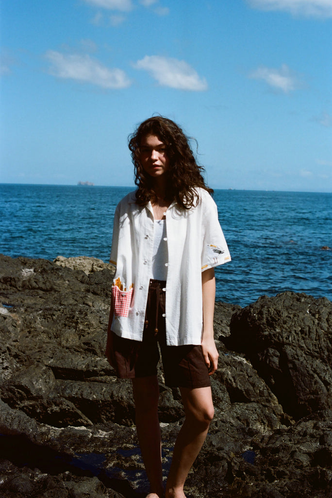 Woman standing on rocky shoreline wearing a white button-down shirt with embroidered fries in a red gingham pocket and fry details on the sleeves.