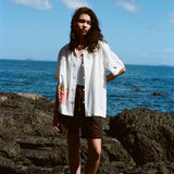 Woman standing on rocks by the ocean wearing a white short-sleeve button-down shirt with French fry embroidery on the red gingham pocket and a seagull embroidered on the sleeve. Shirt is styled open over a white top and paired with dark brown shorts.