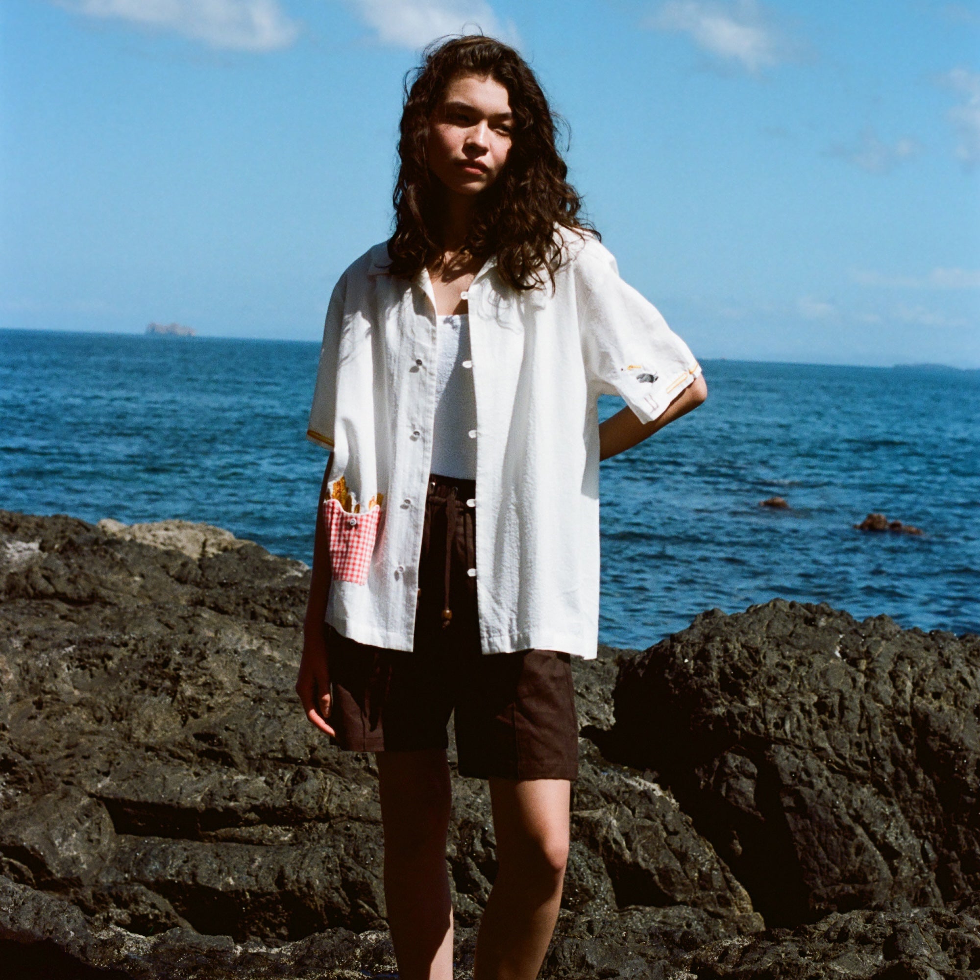 Woman standing on rocks by the ocean wearing a white short-sleeve button-down shirt with French fry embroidery on the red gingham pocket and a seagull embroidered on the sleeve. Shirt is styled open over a white top and paired with dark brown shorts.