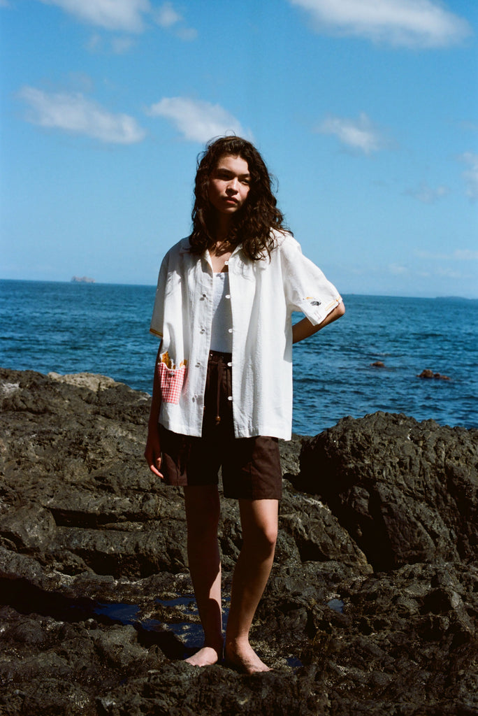 Woman standing on rocks by the ocean wearing a white short-sleeve button-down shirt with French fry embroidery on the red gingham pocket and a seagull embroidered on the sleeve. Shirt is styled open over a white top and paired with dark brown shorts.