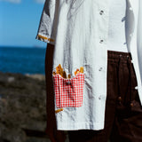 Close-up of a white short-sleeve shirt featuring a red gingham pocket embroidered with golden French fries, worn with brown drawstring shorts.
