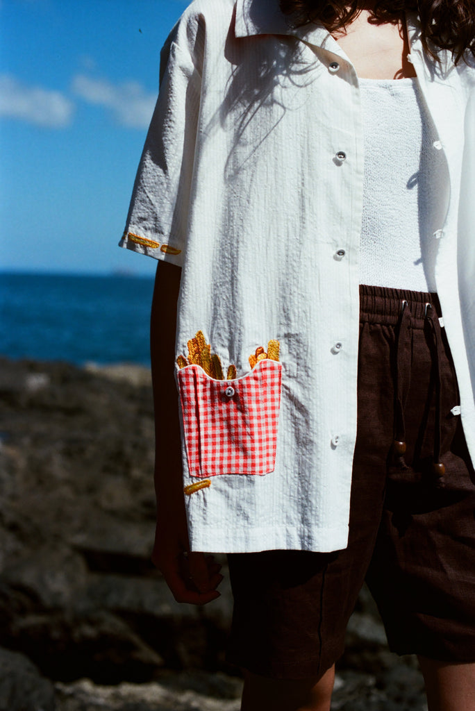 Close-up of a white short-sleeve shirt featuring a red gingham pocket embroidered with golden French fries, worn with brown drawstring shorts.