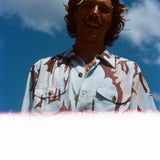 Man with brown hair smiling down at the camera, wearing the abstract blue and brown patterned 'Mary Ball x Tombolo' shirt. 