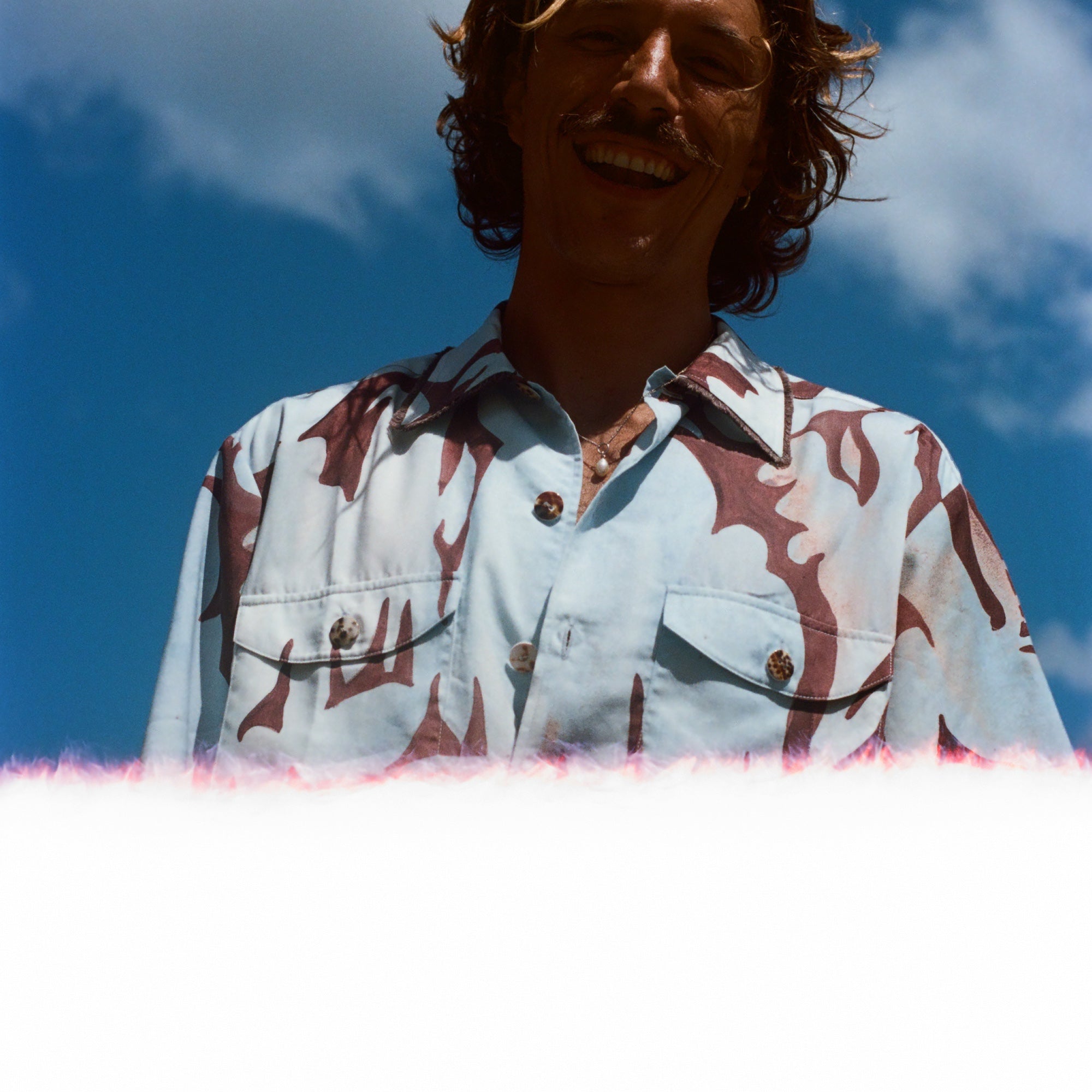 Man with brown hair smiling down at the camera, wearing the abstract blue and brown patterned 'Mary Ball x Tombolo' shirt. 