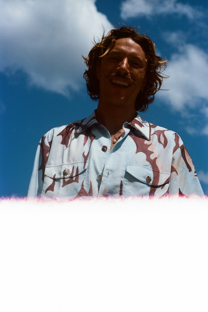 Man with brown hair smiling down at the camera, wearing the abstract blue and brown patterned 'Mary Ball x Tombolo' shirt. 