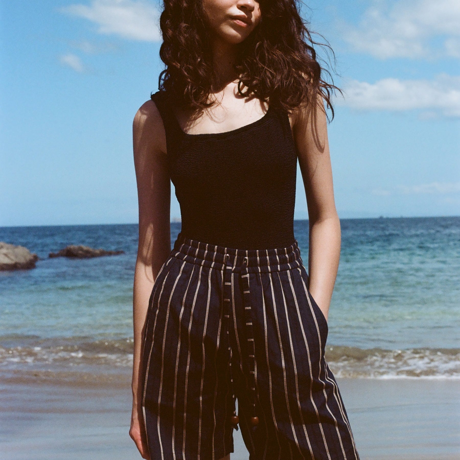 Woman standing on beach, wearing the Easy Going Shorts in Navy Jacquard, with a blank tank top, and one hand in her pocket.