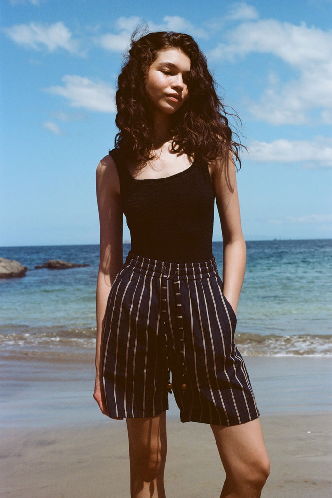 Woman standing on beach, wearing the Easy Going Shorts in Navy Jacquard, with a blank tank top, and one hand in her pocket.