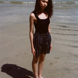 Woman standing on beach, wearing the Easy Going Shorts in Navy Jacquard, with a blank tank top and white sandals. 