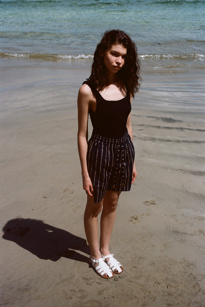 Woman standing on beach, wearing the Easy Going Shorts in Navy Jacquard, with a blank tank top and white sandals. 