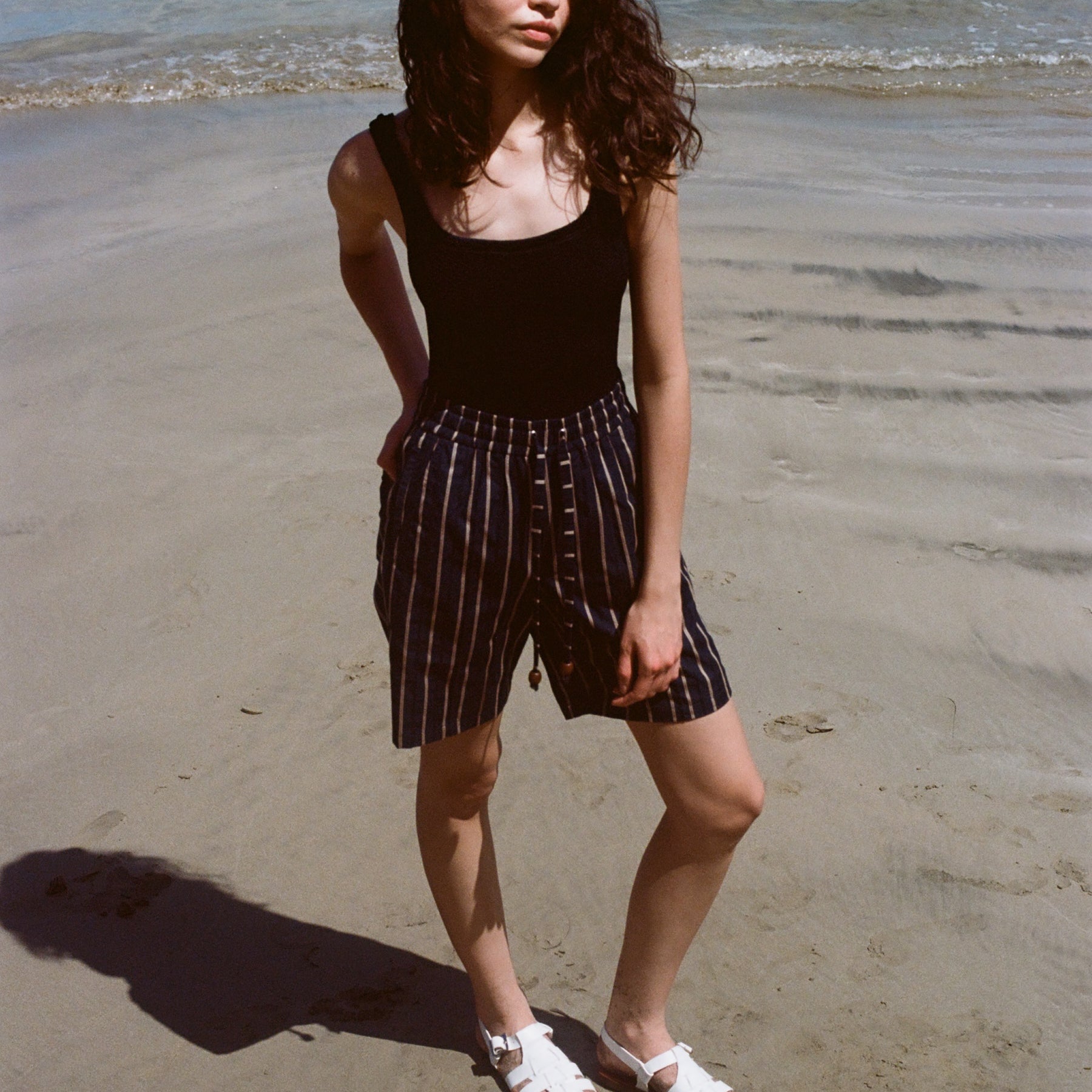 Woman standing on beach, wearing the Easy Going Shorts in Navy Jacquard, with a blank tank top, and one hand in her pocket. She has white sandals on.