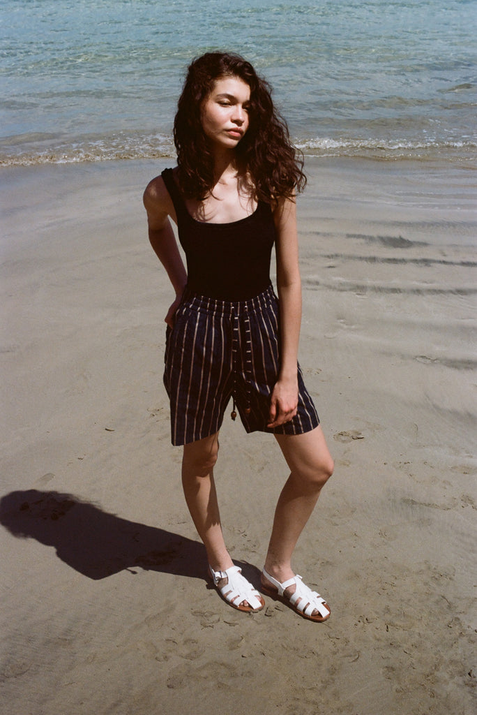 Woman standing on beach, wearing the Easy Going Shorts in Navy Jacquard, with a blank tank top, and one hand in her pocket. She has white sandals on.
