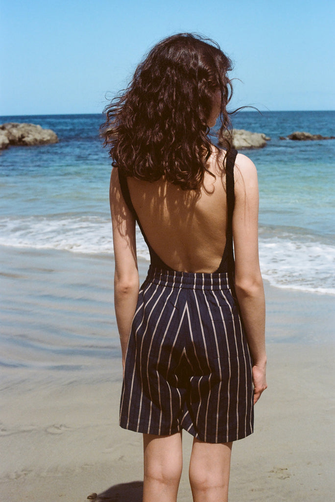 Woman standing on a beach with her back to us. She is wearing a black tank top and our Navy Easy Going Shorts with beige stripes. 