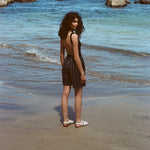 Woman standing on beach, wearing the Easy Going Shorts in Navy Jacquard, with a blank tank top, and white sandals. She is facing the ocean but looks back at the camera. 