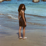 Woman standing on beach, wearing the Easy Going Shorts in Navy Jacquard, with a blank tank top, and white sandals. She is facing the ocean but looks back at the camera. 