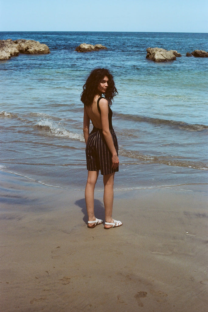 Woman standing on beach, wearing the Easy Going Shorts in Navy Jacquard, with a blank tank top, and white sandals. She is facing the ocean but looks back at the camera. 