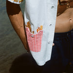 Close-up of a man wearing a white short-sleeve shirt with a red gingham pocket embroidered with French fries, standing near the water.