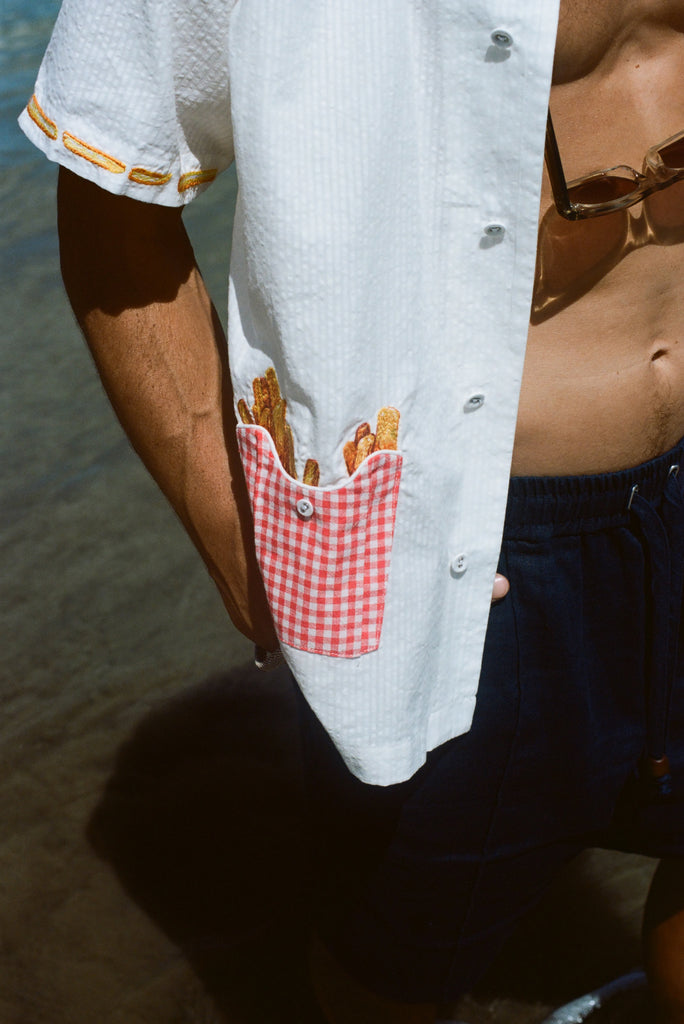 Close-up of a man wearing a white short-sleeve shirt with a red gingham pocket embroidered with French fries, standing near the water.