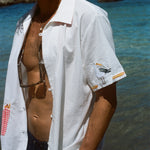 Close-up of a man wearing a white shirt with a red gingham pocket of embroidered French fries and a seagull stitched on the sleeve, standing near the ocean.