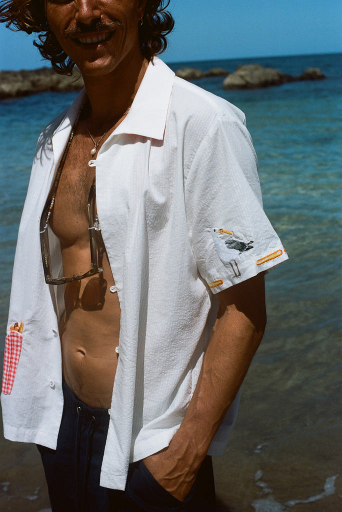 Close-up of a man wearing a white shirt with a red gingham pocket of embroidered French fries and a seagull stitched on the sleeve, standing near the ocean.