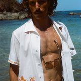 Man standing in front of the ocean wearing a white short-sleeve shirt with French fry embroidery on the red gingham pocket and seagull embroidery on the sleeve. The shirt is unbuttoned.