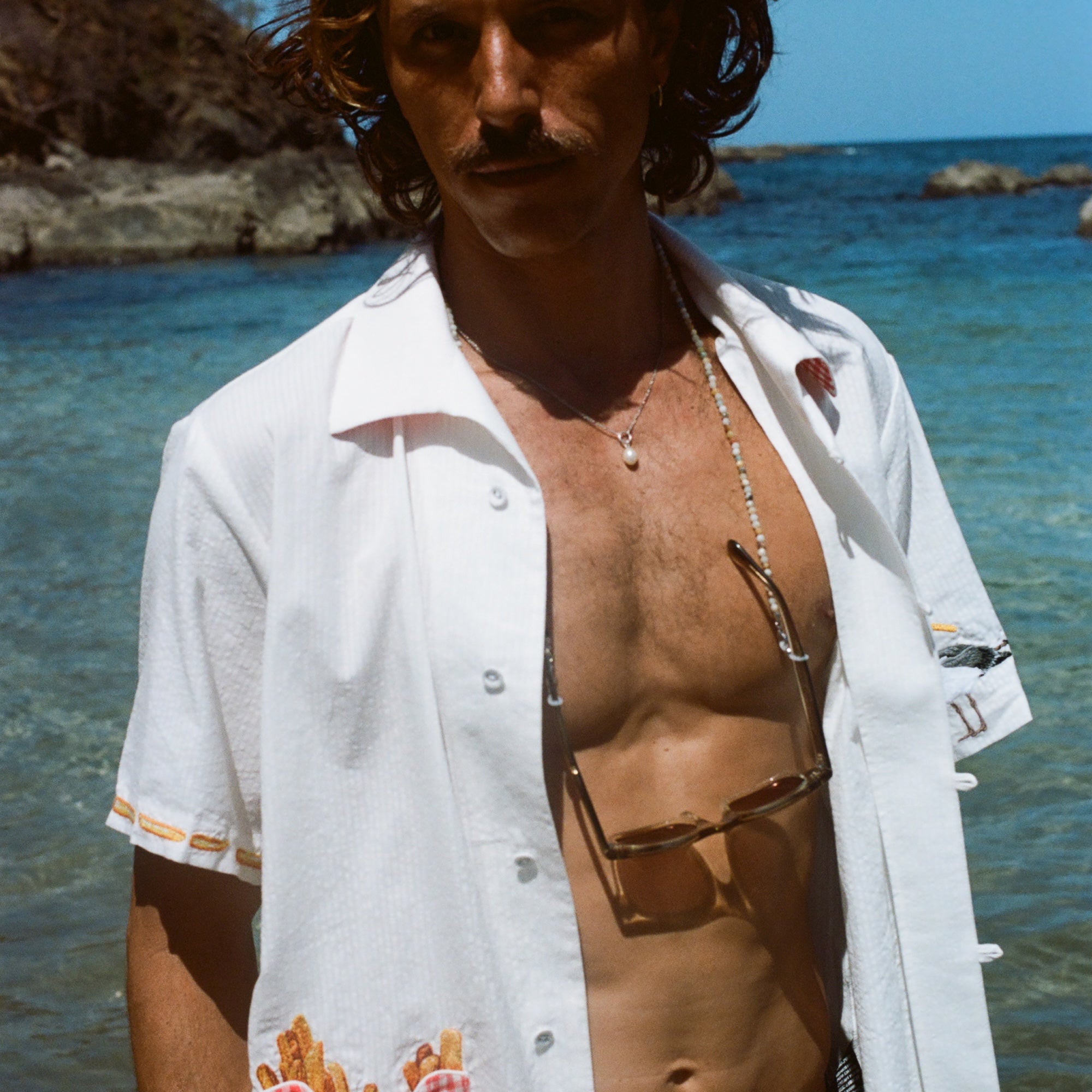 Man standing in front of the ocean wearing a white short-sleeve shirt with French fry embroidery on the red gingham pocket and seagull embroidery on the sleeve. The shirt is unbuttoned.