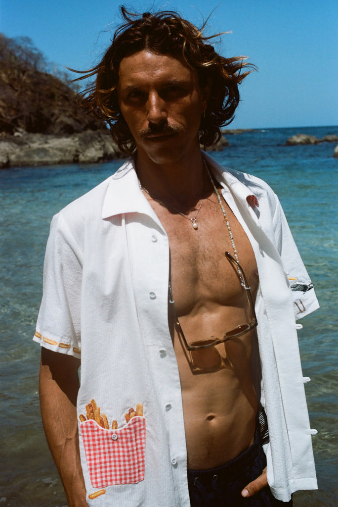 Man standing in front of the ocean wearing a white short-sleeve shirt with French fry embroidery on the red gingham pocket and seagull embroidery on the sleeve. The shirt is unbuttoned.