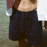 Close-up image of a person standing in ocean water wearing navy linen shorts with a drawstring and wooden bead accents. The model is also wearing a white shirt with a red gingham pocket graphic featuring fries.