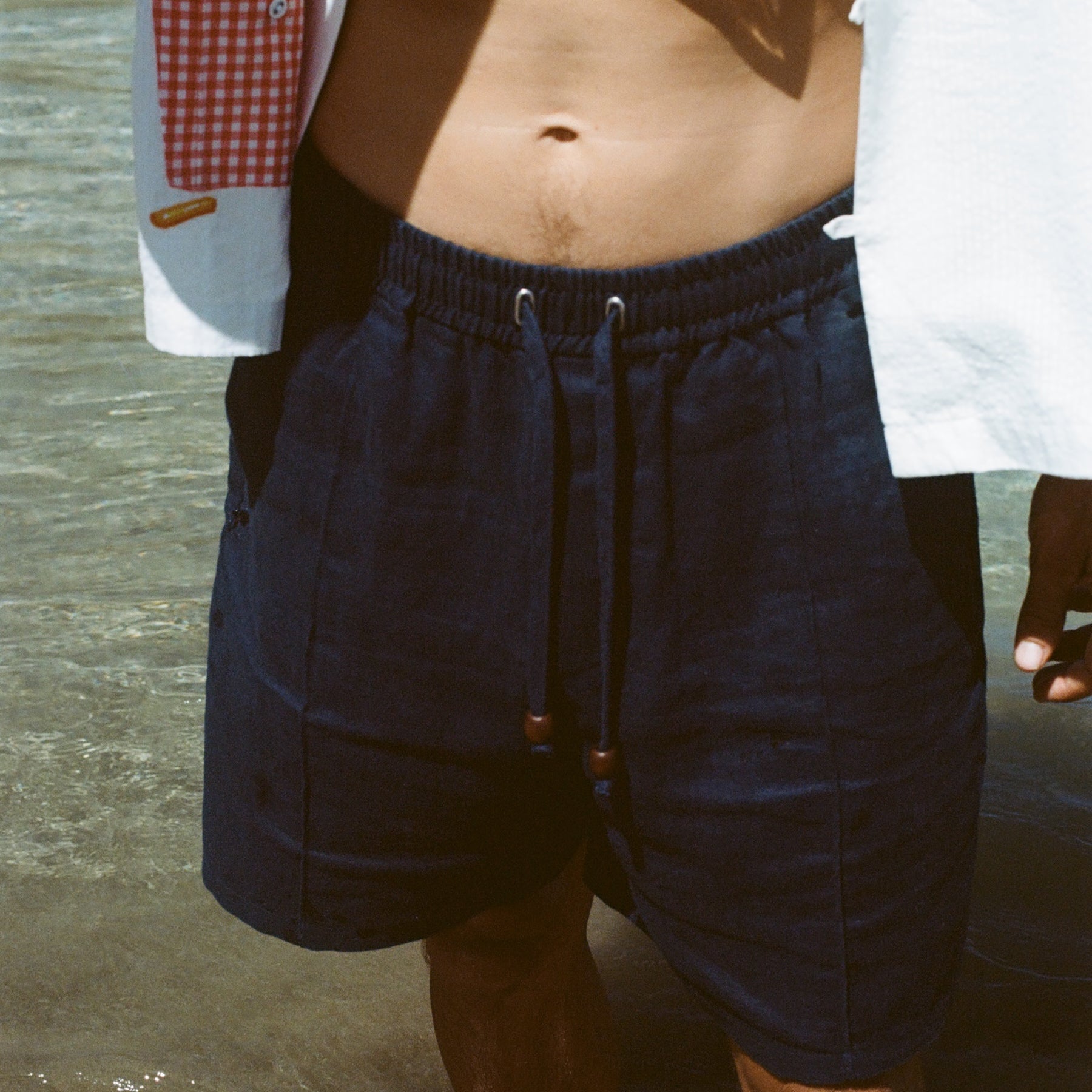 Close-up image of a person standing in ocean water wearing navy linen shorts with a drawstring and wooden bead accents. The model is also wearing a white shirt with a red gingham pocket graphic featuring fries.
