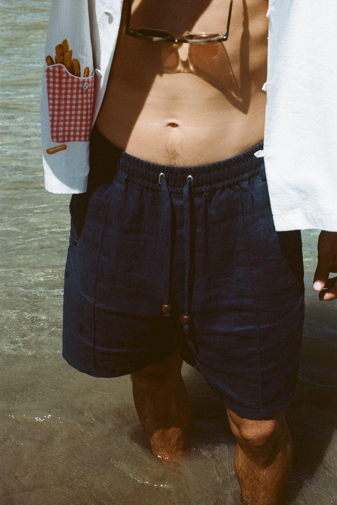 Close-up image of a person standing in ocean water wearing navy linen shorts with a drawstring and wooden bead accents. The model is also wearing a white shirt with a red gingham pocket graphic featuring fries.
