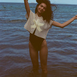 Woman playfully throwing her arms up while standing ankle deep in the ocean, with the 'Starfish Strut' Hand Towel Top over a black one-piece swimsuit.