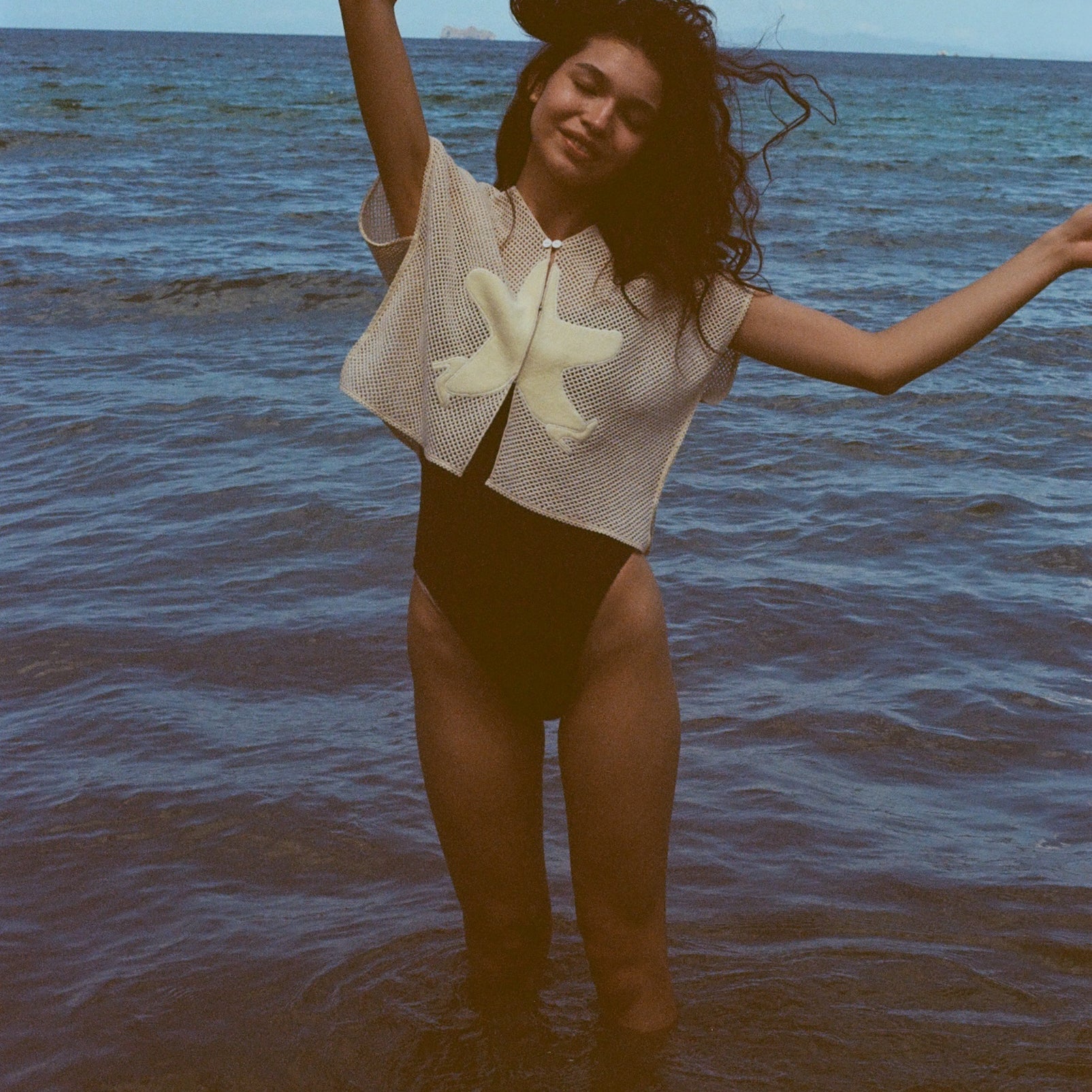 Woman playfully throwing her arms up while standing ankle deep in the ocean, with the 'Starfish Strut' Hand Towel Top over a black one-piece swimsuit.