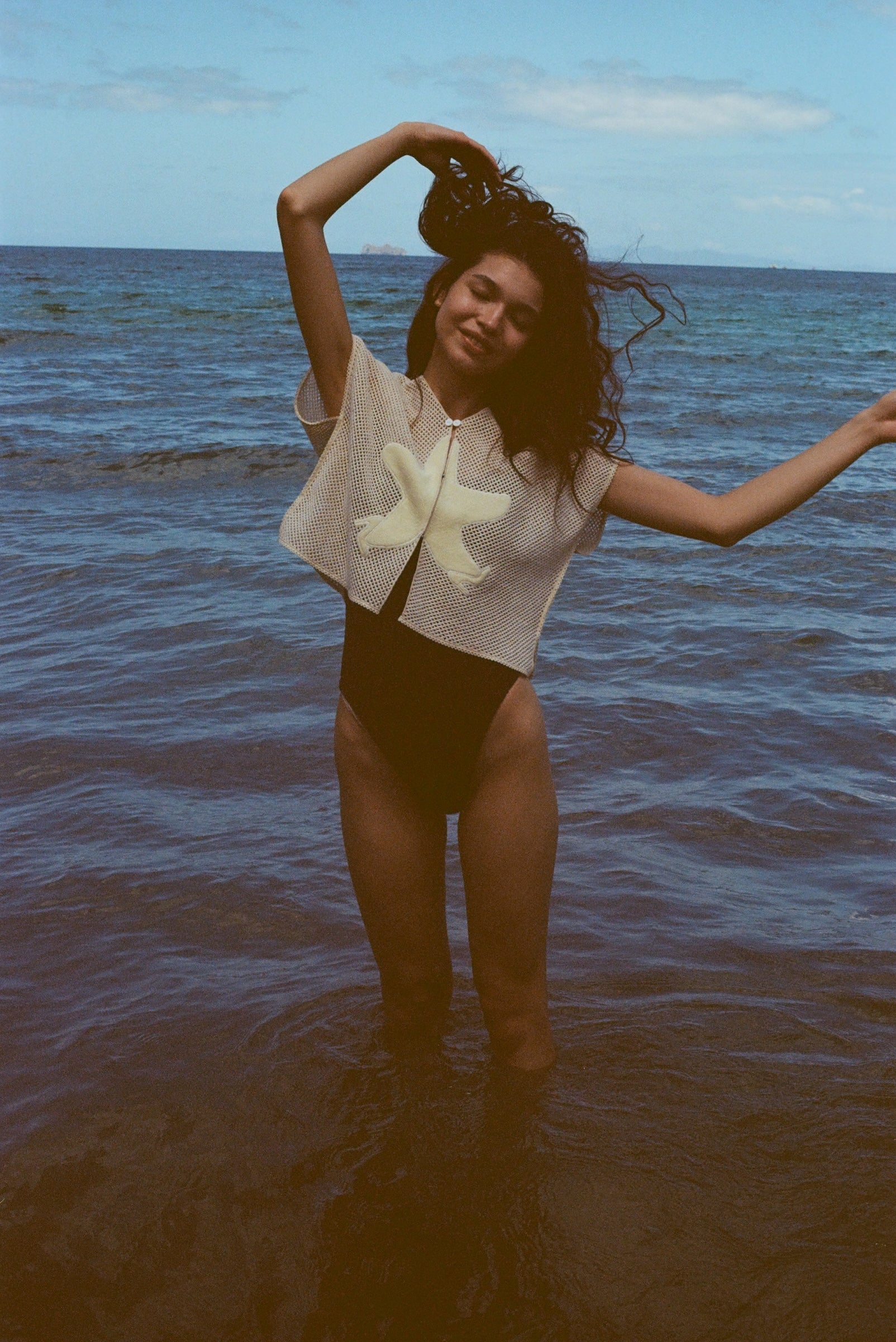 Woman playfully throwing her arms up while standing ankle deep in the ocean, with the 'Starfish Strut' Hand Towel Top over a black one-piece swimsuit.