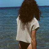 A woman stands waist-deep in the ocean, facing away from the camera. She wears  the 'Starfish Strut' Hand Towel Top over a black swimsuit bottom, with dark curly hair cascading down her back. 