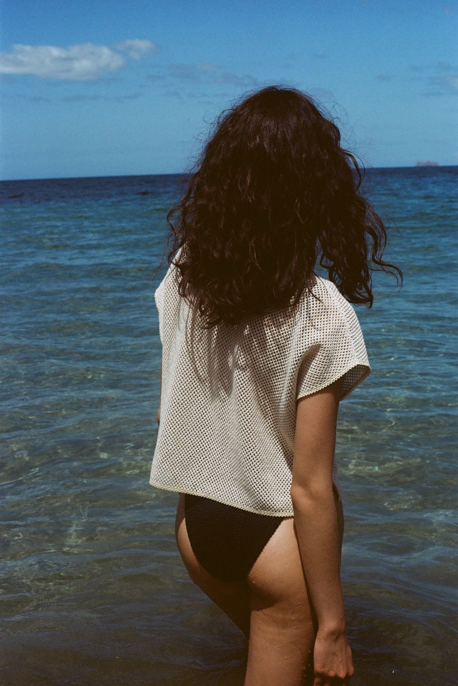 A woman stands waist-deep in the ocean, facing away from the camera. She wears  the 'Starfish Strut' Hand Towel Top over a black swimsuit bottom, with dark curly hair cascading down her back. 