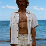 Brunette man standing on beach while wearing the sand linen Easy Going Shorts with our featherweight Resort Shirt fully unbuttoned. 