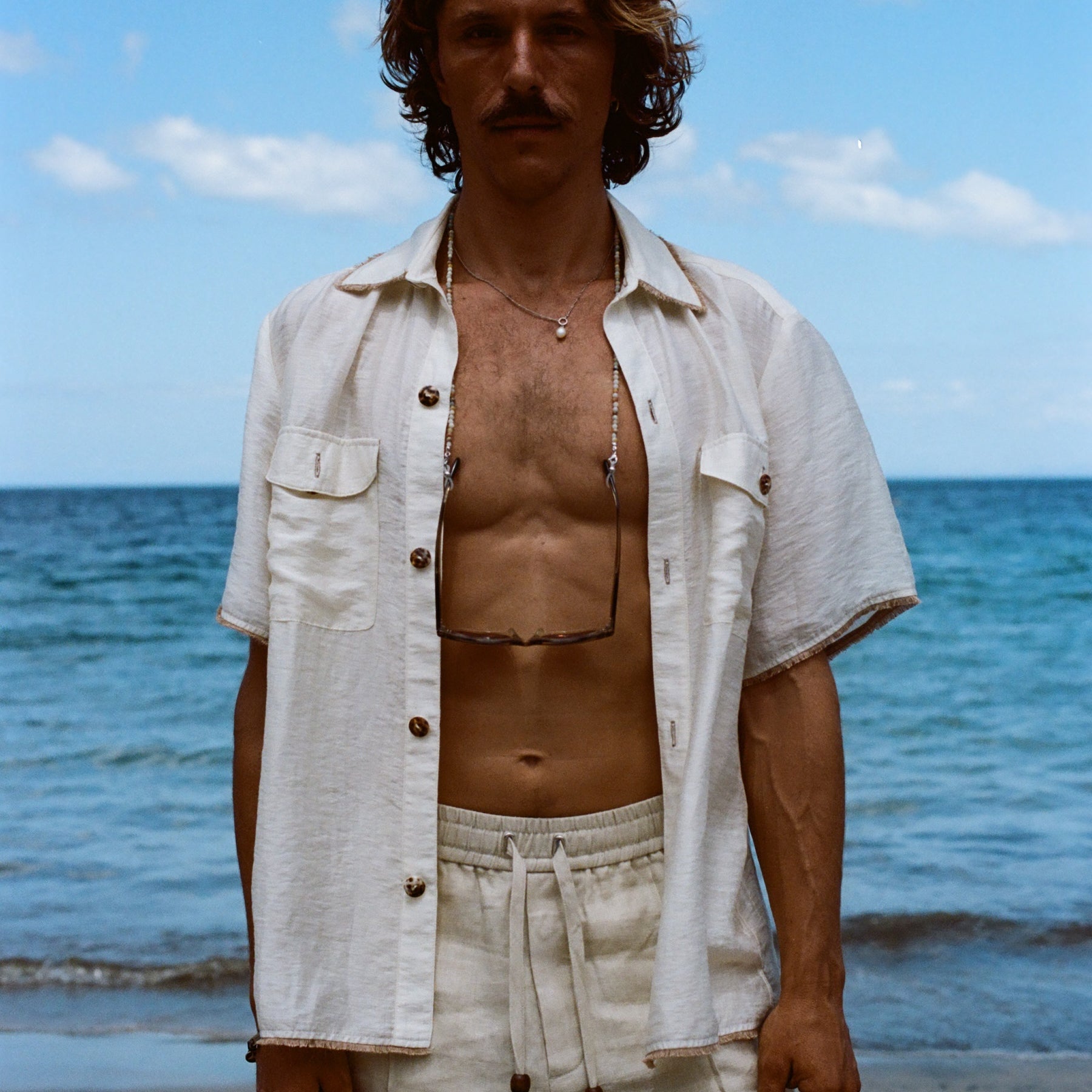 Man at the beach facing the camera wearing a button down white Tombolo shirt and beige shorts 