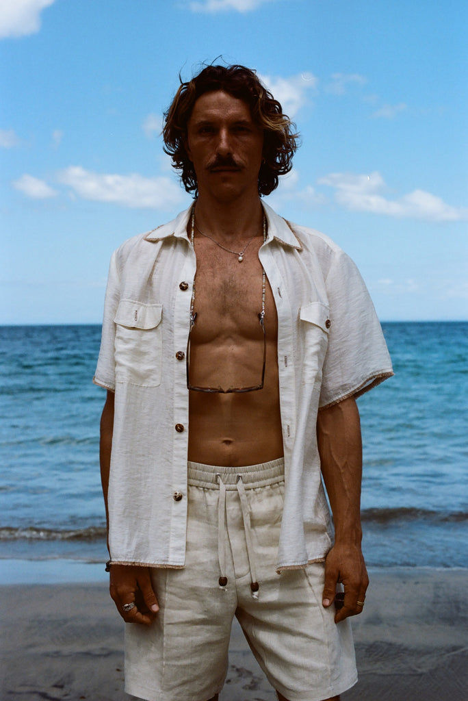 Man at the beach facing the camera wearing a button down white Tombolo shirt and beige shorts 