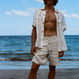 Brunette man standing on beach while wearing the sand linen Easy Going Shorts with our featherweight Resort Shirt fully unbuttoned. 