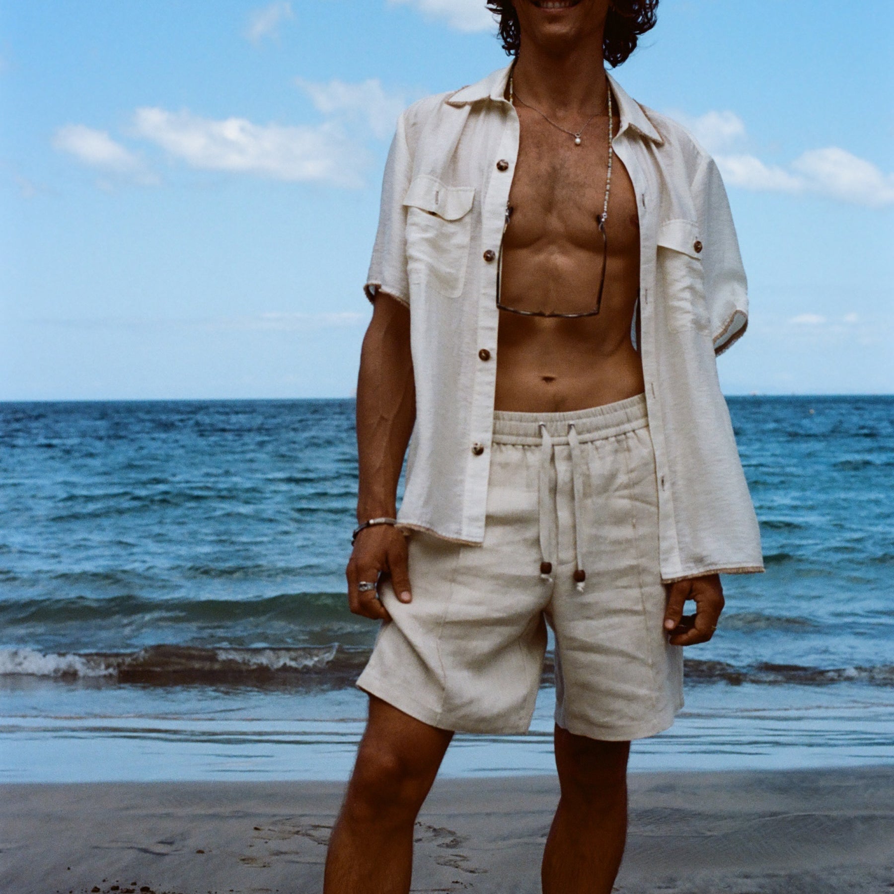 Brunette man standing on beach while wearing the sand linen Easy Going Shorts with our featherweight Resort Shirt fully unbuttoned. 