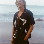 Male model gazing up on the beach wearing the Black Cabana Shirt with one hand in his pocket. He wears the shirt unbuttoned and relaxed. 