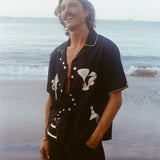 Male model gazing up on the beach wearing the Black Cabana Shirt with one hand in his pocket. He wears the shirt unbuttoned and relaxed. 
