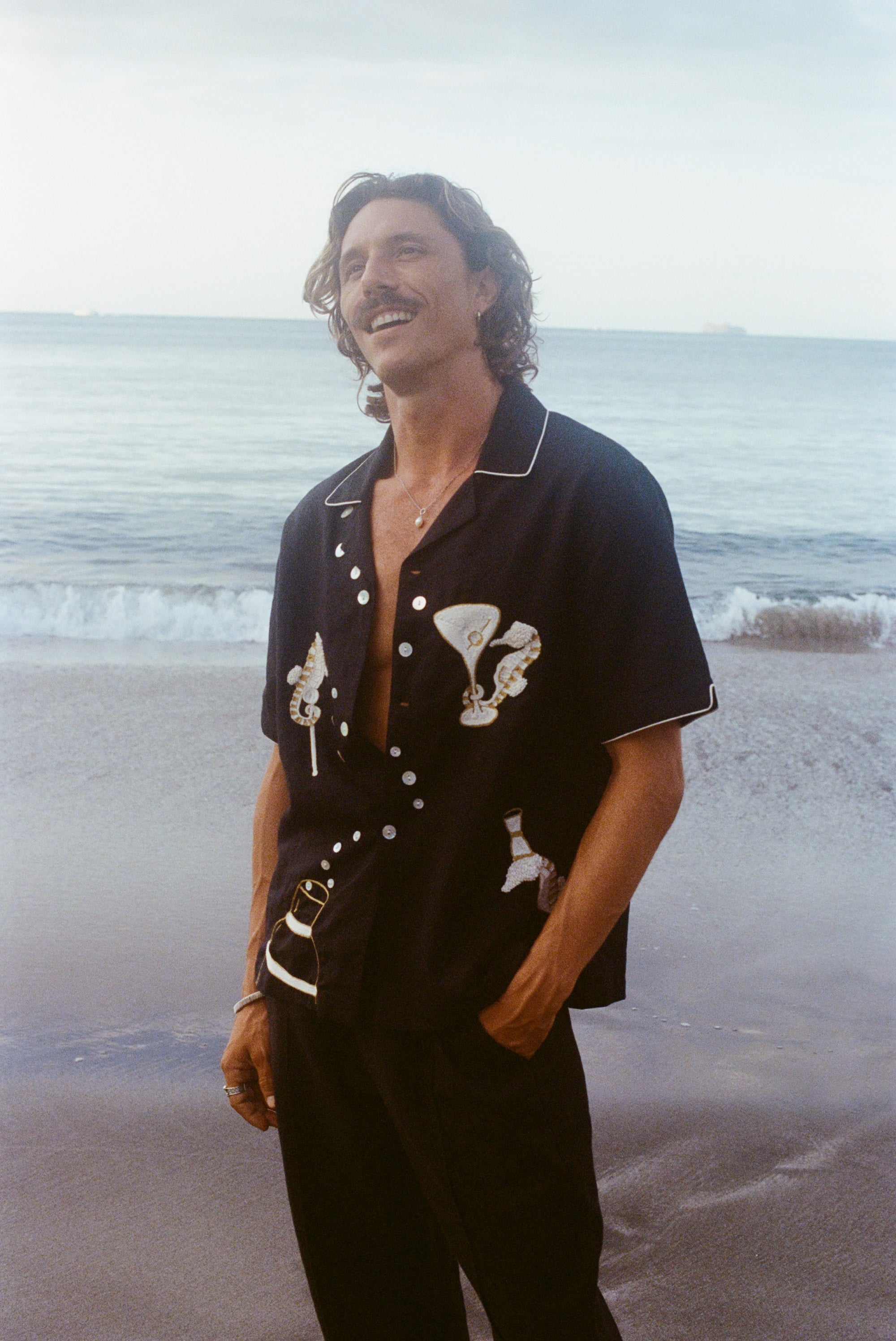 Male model gazing up on the beach wearing the Black Cabana Shirt with one hand in his pocket. He wears the shirt unbuttoned and relaxed. 