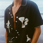 A smiling man wearing 'Tip Your Bartenders!' Cabana fully unbuttoned standing on the beach. 