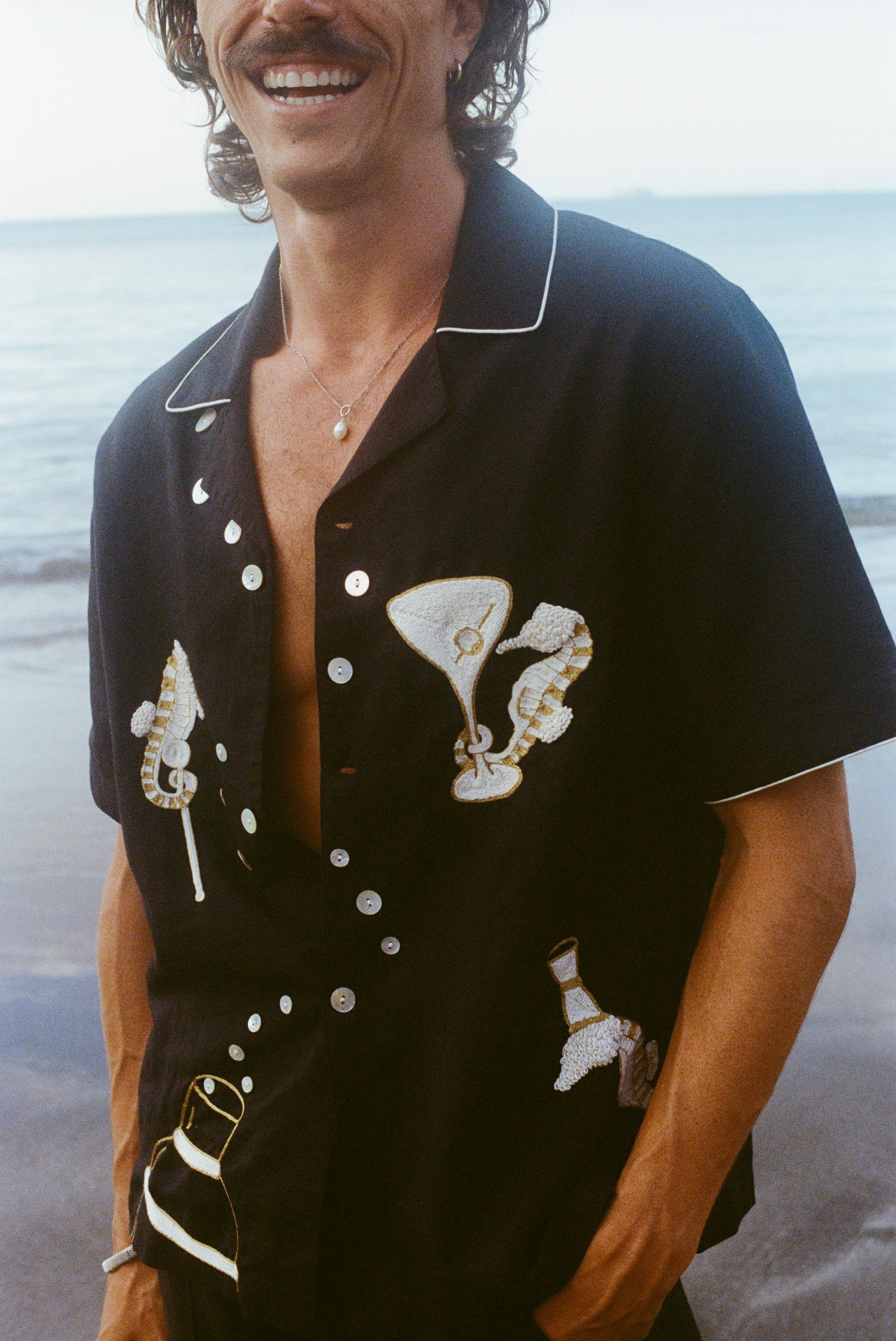 A smiling man wearing 'Tip Your Bartenders!' Cabana fully unbuttoned standing on the beach. 