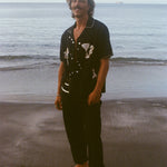 Male model pictured standing barefoot on beach, smiling at camera and wearing the Black button down shirt open with black pants. 