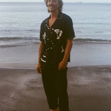 Male model pictured standing barefoot on beach, smiling at camera and wearing the Black button down shirt open with black pants. 