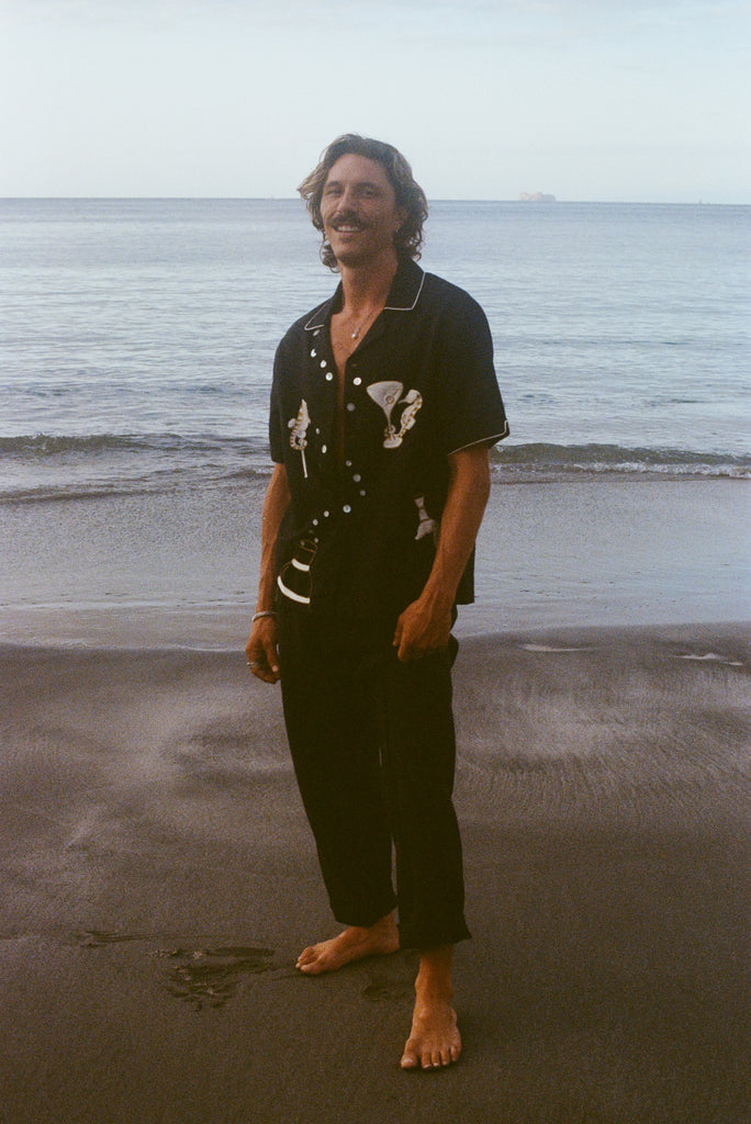 Male model pictured standing barefoot on beach, smiling at camera and wearing the Black button down shirt open with black pants. 