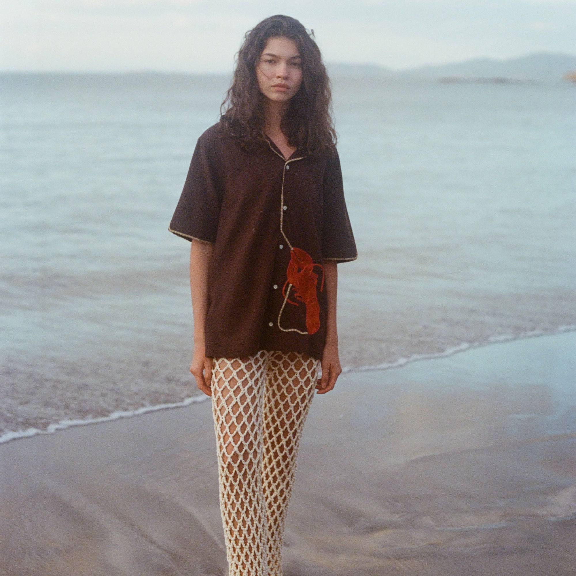 Woman standing on the beach wearing cream open knit pants and a Tombolo Brown Button Down ('Bisque Averse' Cabana), gazing directly into camera on a cloudy day.