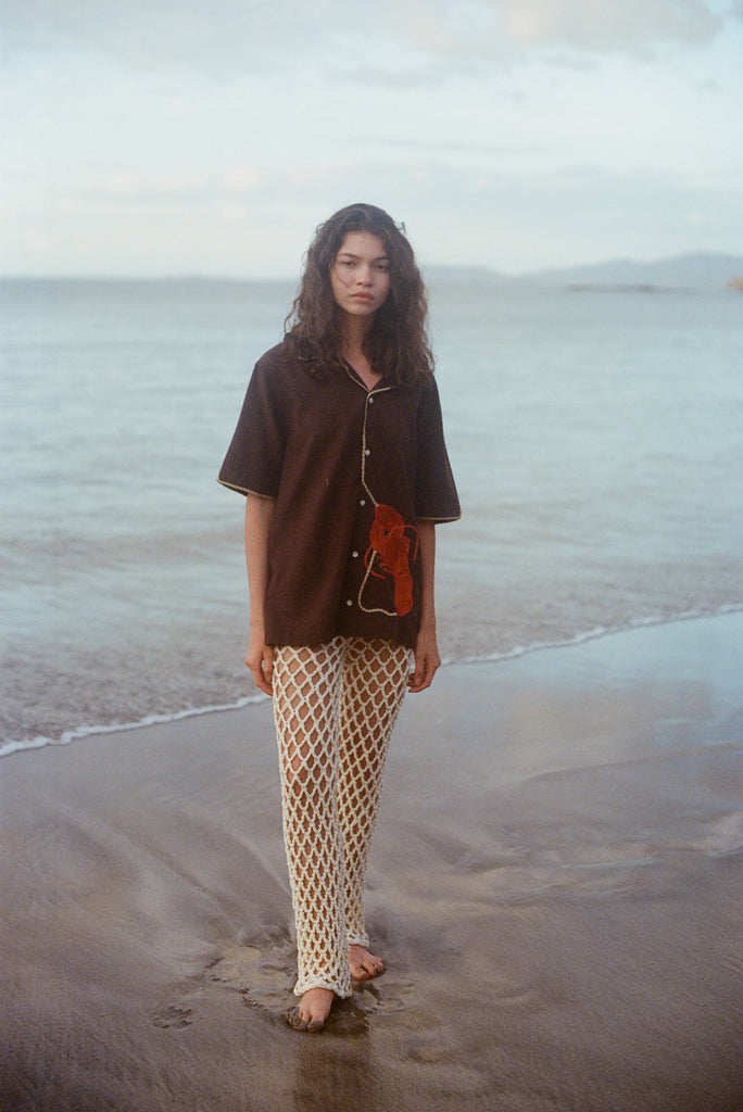 Woman standing on the beach wearing cream open knit pants and a Tombolo Brown Button Down ('Bisque Averse' Cabana), gazing directly into camera on a cloudy day.