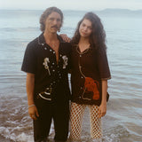 Two models stand ankle-deep in ocean water at sunset. The male model wears a black short-sleeve button-down shirt embroidered with two seahorses ('Tip your Bartenders' Cabana), paired with matching black trousers. The female model wears a brown short-sleeve shirt with beige piping and a large red lobster applique on the lower front ("Bisque Averse"), paired with white open-knit pants. They stand close together with the woman's hand resting gently on the man's shoulder.